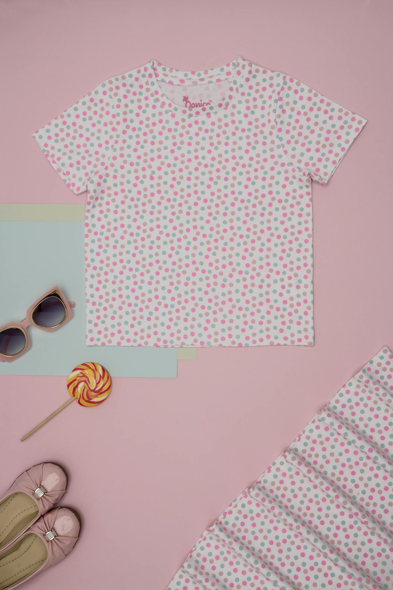 Kids' fashion flat lay with polka dot shirt, sunglasses, shoes, and lollipop on a pink background.